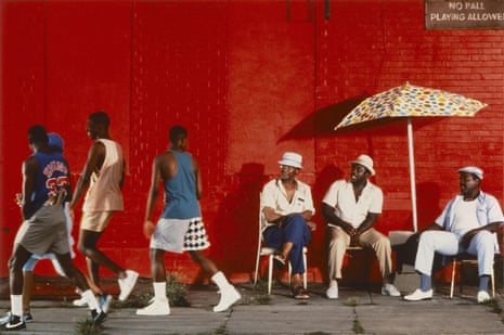 A man walks past three men seated under a polka dot umbrella against a bright red brick wall