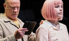 A man in a boiler suit with a computer tablet in his right hand, apparently a technician, has his hand at the back of a woman with pink hair in a bob style and wearing a pink uniform with a logo saying "De Santo". She appears to be a robot being programmed by him, by pressing keys/buttons on her back.