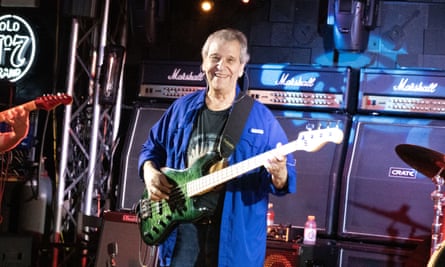 An older man with a bass guitar on his shoulders in front of three Marshall stacks.