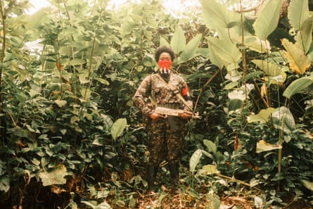 A fighter in combat gear with a red and black bandana covering their nose and mouth poses with their rifle surrounded by jungle foliage