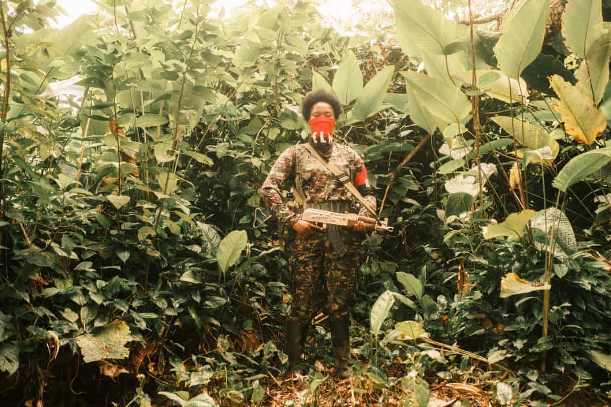 A fighter in combat gear with a red and black bandana covering their nose and mouth poses with their rifle surrounded by jungle foliage