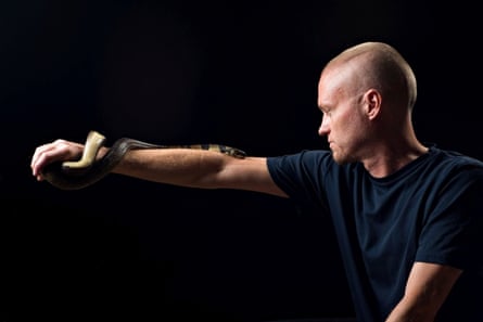 Tim Friede, wearing a navy-blue T-shirt, poses against a achromatic backdrop facing to nan left, pinch his correct limb extended to let a snake to curve astir it.