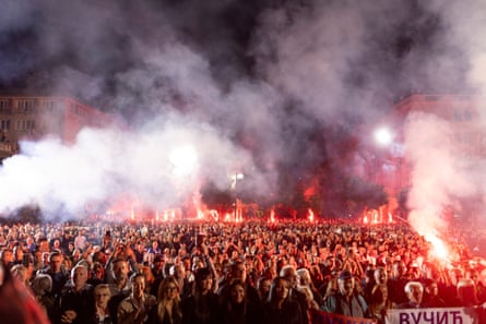 Smoke and the flames from flares cast a red glow over people crowded in a square at night