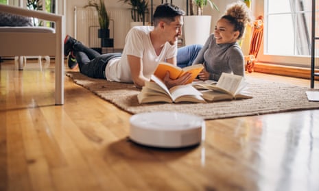 A robot vacuum cleaner in the foreground of a photograph of a young couple lying on the floor together.