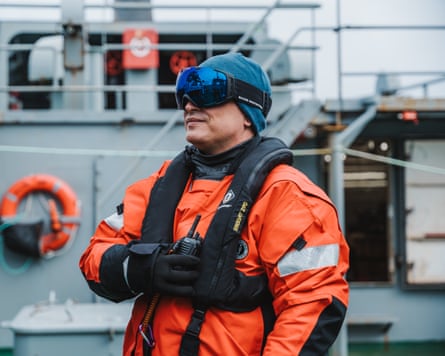 A man in an orange suit and goggles stands on a boat deck