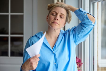A woman with eyes closed, probably in her late 40s, in a sky blue shirt facing the camera, holding her hair up and fanning herself with a sheet of white paper –in a room with windows behind and to the side of her