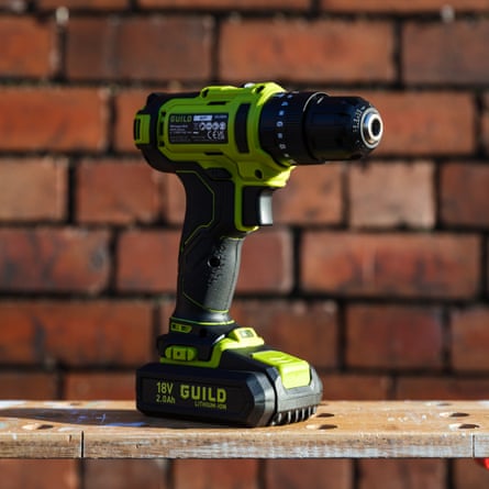 The Guild Cordless impact drill is standing upright in the sunlight against a red brick wall. The drill is neon yellow and black.