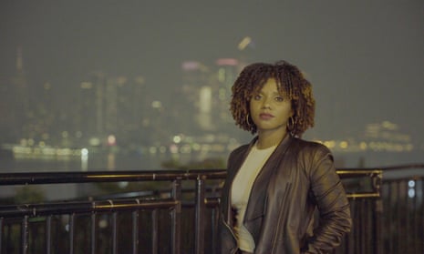 A young Black woman with a curly bob stands outside in the dark with a city skyline beyond her, wearing a white T-shirt and black leather jacket.