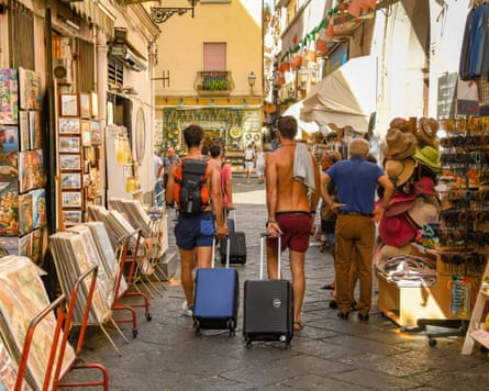 Two shirtless tourists pulling suitcases past souvenir shops in the old town in Sorrento