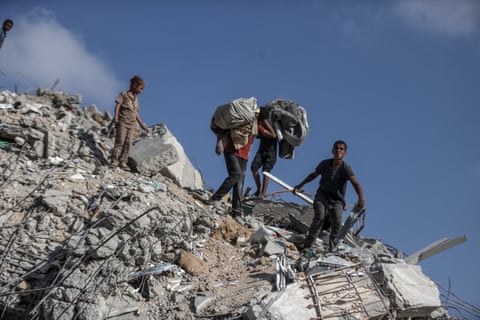 مردان و کودکان از آوار بتنی و میلگرد بالا میروند و کیسههایی را روی سر و شانههایشان نگه داشتهاند.