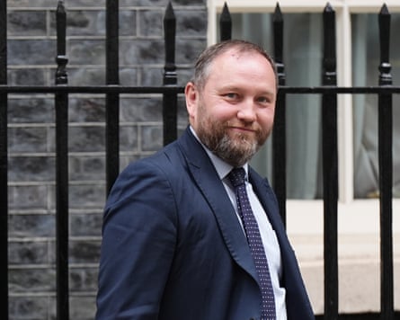 he is round-faced with short, light-brown receding hair and a trimmed beard, and wears a dark jacket and tie. There are black iron railings behind him.