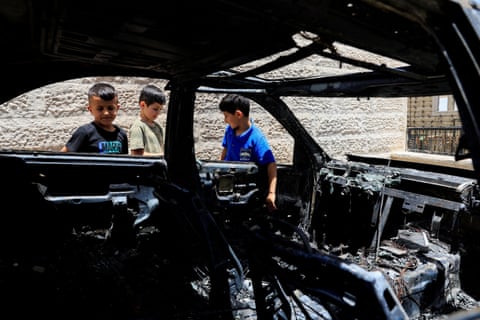 Three young boys stand around a burned-out car, photographed through its blackened, glassless windows.