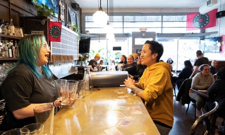 A woman in a yellow hoodie stands at a bar talking to a woman behind the bar holding beer glasses.