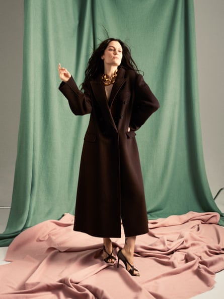 Jessie J standing in front of a piece of draped green material and standing on a pale-orange piece of material, wearing a full-length dark-brown coat, dark-brown strappy shoes and a chunky gold necklace, her right arm held out bent at the elbow and her left hand on her waist