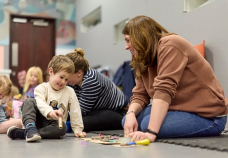 A boy sitting on the floor smiles as he plays with craft materials while a woman sitting next to him looks on