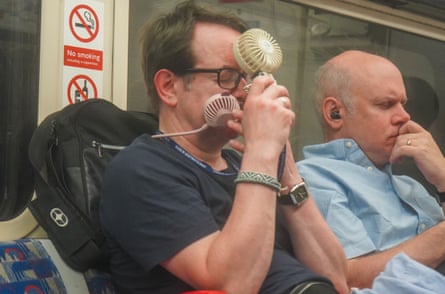 A man holds two handheld fans to his face as he tries to keep cool in his seat on the tube