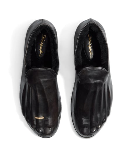 A pair of black shoes with the outline of fingers including the nails on the top of the shoe, with a gold ring on the second ‘finger’ of the right-hand shoe