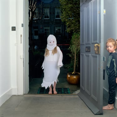 A child dressed in a skeleton top stands hidden behind an open front door as another child in a white dress and white balaclava-like hood steps over the threshold