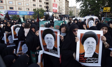 Iranian women hold posters depicting the president during a mourning ceremony in Tehran, 20 May 2024: they are all dressed in black and have their heads covered. The crowd stretches across a street and shops can be seen in the back