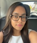 Ria appears to be sitting in a car. She has long, straight dark brown hair and is wearing large black-framed glasses and a white vest top.
