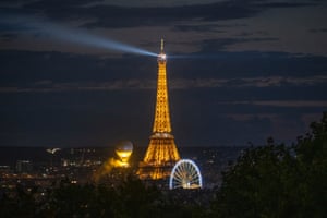 A searchlight shines out into the night sky from the top of the illuminated Eiffel Tower as the Paris 2024 Olympic cauldron, looking like a giant hot-air balloon, hovers low over the ground. In the foreground, the illuminated Paris ferris wheel