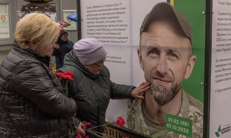 Mariia Kurbet, holding red flowers in one hand, touches a photographic portrait of her son, Vasyl Kurbett, in military uniform, with the dates 01.02.1981 and 07.10.2022