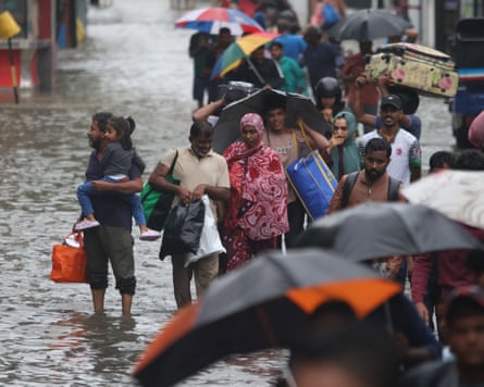 Crowds of people wading through ankle-deep water carrying bags and umbrellas