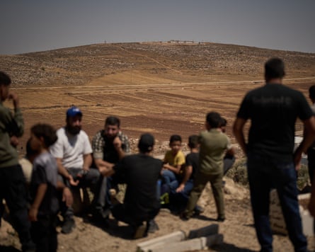 A small group of Palestinians on an embankment look out at an Israeli settlement on the next hill