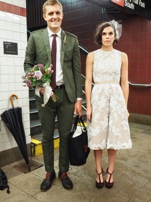A man in a suit and a woman in a lacy white knee-length jumpsuit stand in the New York subway