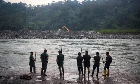 Sete homens carregando lanças estão perto de um rio olhando para uma escavadeira mecânica na margem oposta