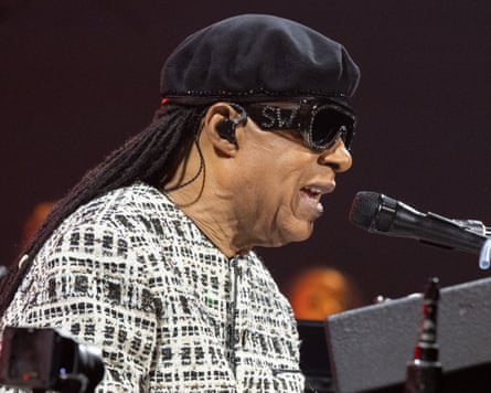 Stevie Wonder, in a cap and sunglasses, sings into a microphone while playing a keyboard