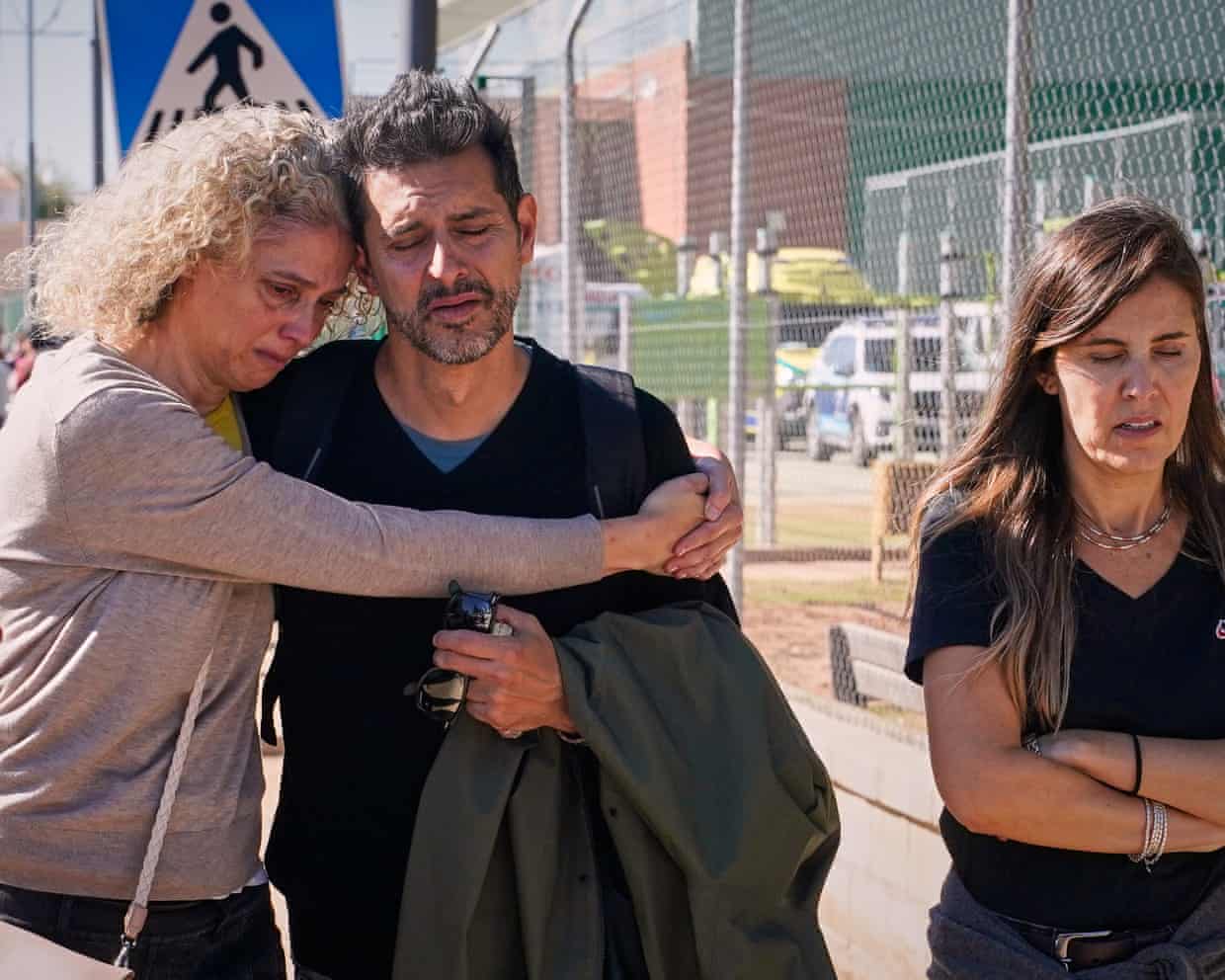 A woman with curly, blond hair wearing a beige top weeps as she holds a grey-haired man in a black V-neck top and rests her head on his. Another woman walks beside them, holding herself and looking traumatised