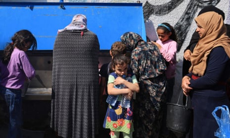 A girl holds two water bottles and people fill up bottles and buckets from a water station behind her