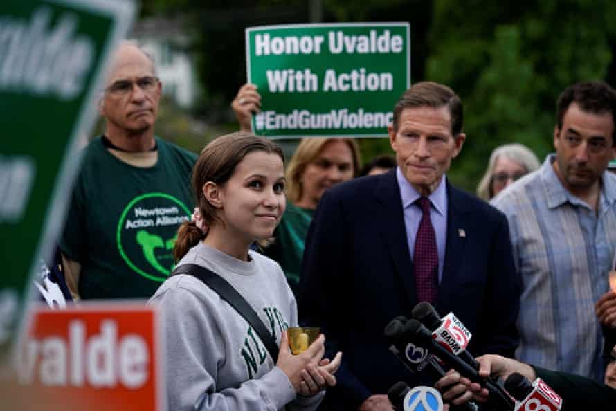 A woman stands among a group of protesters, talking to the media who are holding microphones. A sign in the background reads ‘Honor Uvalde with action. #EndGunViolence’.