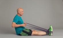 A bald man in his sixties (Phil Daoust), wearing a T-shirt, shorts and trainers, sitting on the floor pulling on resistance bands around his feet