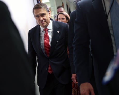 Attorney General nominee Todd Blanche in a dark suit and red tie walks through a corridor surrounded by others