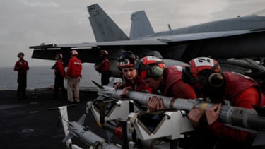 A video screenshot shows US Navy sailors loading munitions onto a fighter jet on the flight deck.