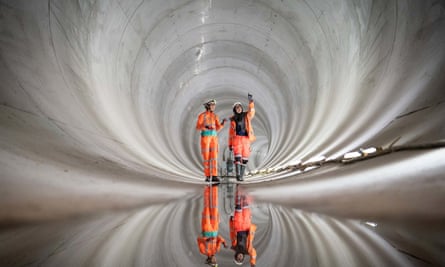 Two workers in hi-vis orange workwear walk a long al long bare concrete tunnel lit from ground level