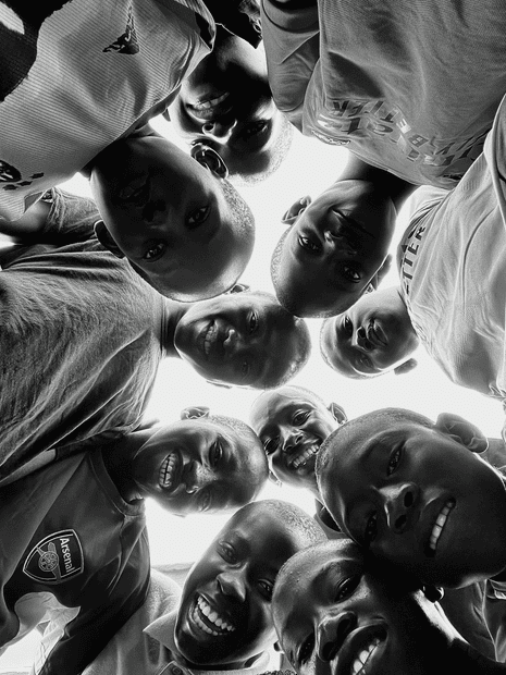 A black and white photo of the faces of young football fans in the Gambia, in a huddle, looking down