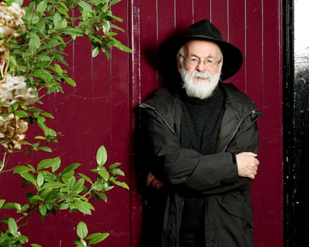 Terry Pratchett stands in front of a dark red wooden door beside a tall green-leaved bush; he has a thick white beard and wears a big black hat with wide brim, plus black jumper and anorak.