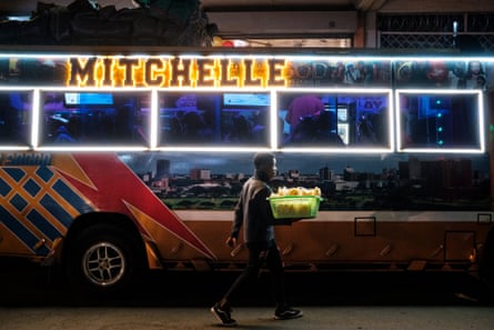 A man carrying a green basket of snacks walks past a matatu decorated with lighting and artwork