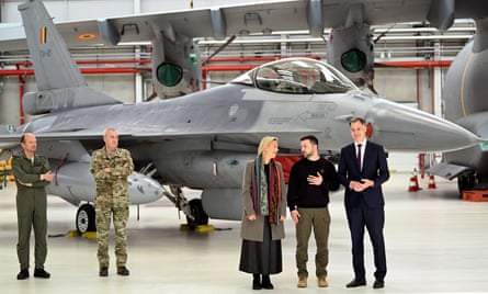 Zelenskiy, De Croo, Dedonder and two other people in military overalls stand in front of an F-16 in an aircraft hangar.