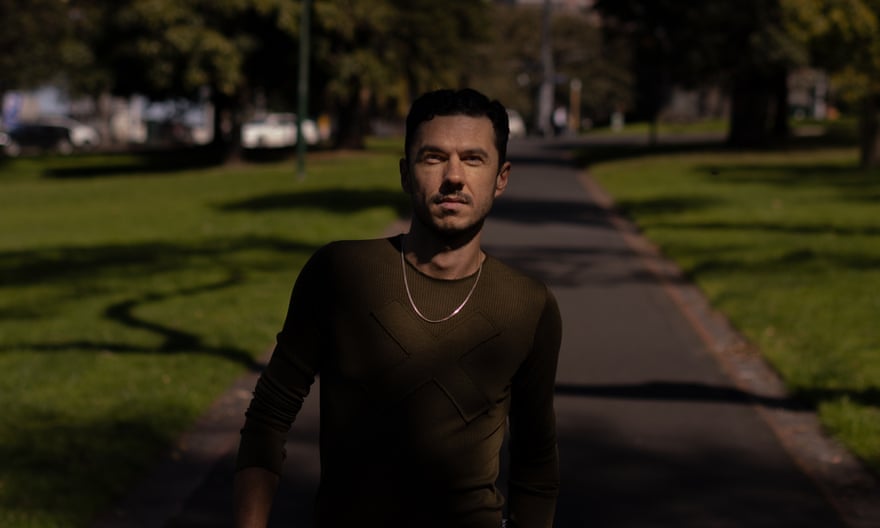 Goran Stolevski walks along a shady path in a park wearing a sweater and neck chain
