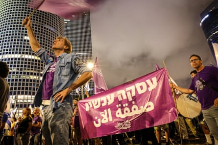 A man waves a purple flag while other protesters hold a banner in a city centre, and another protester holds a drum