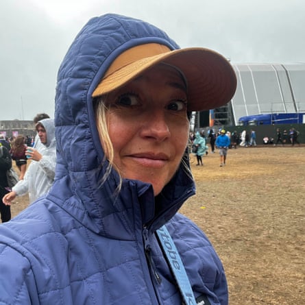 A woman in a hooded Finisterre jacket and visor smiles, with a muddy festival field behind her