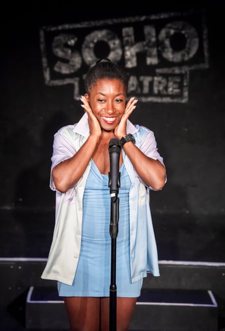 Comedian Sophie Duker smiles, cupping her face behind a stand microphone on stage