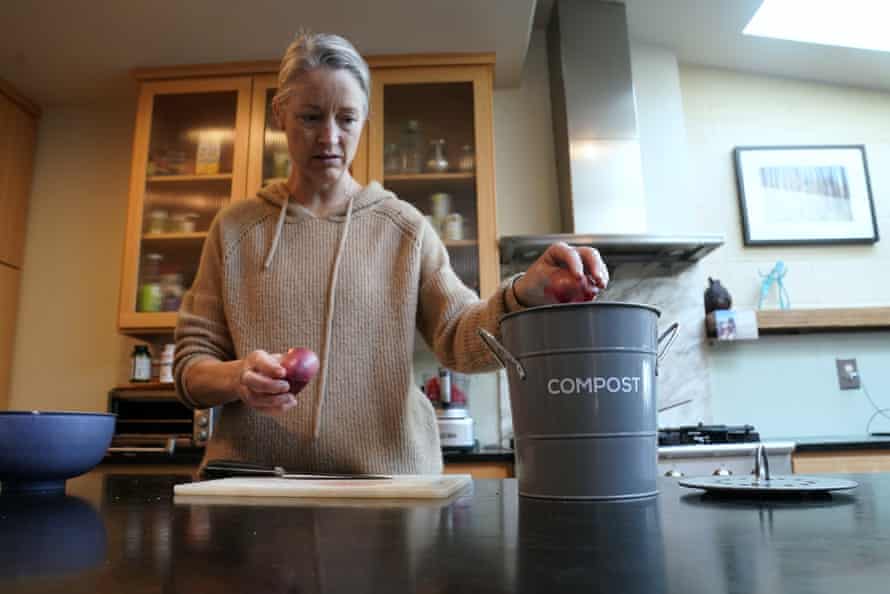 A woman places onion scraps into a gray bucket labeled "compost" on her kitchen counter.