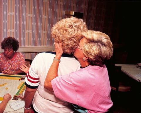 A photograph of a woman sat on another's lap while she whispers in her ear while playing Scrabble