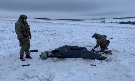 Two members of Ukrainian police, decked in camouflage gear, kneel before a Russian kamikaze unmanned aerial vehicle that lies on the ground of a snowy backdrop. 