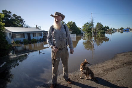 A man with a white beard and wearing a broad-brimmed hat stands on the edge of flood waters with a dog sitting next to his feet. Behind him a house is inundated on a residential street